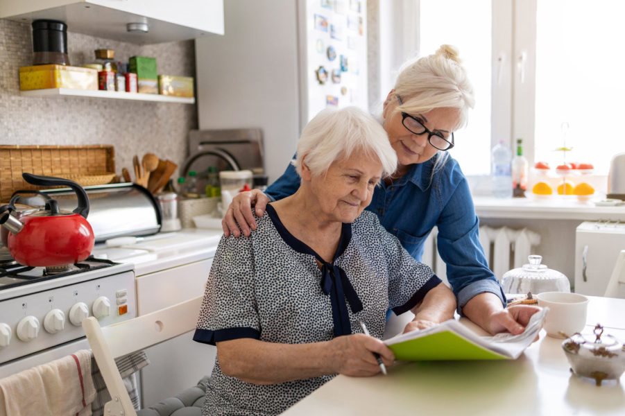 Caregiver daughter helping elderly mother review and sign paperwork together at a home kitchen table