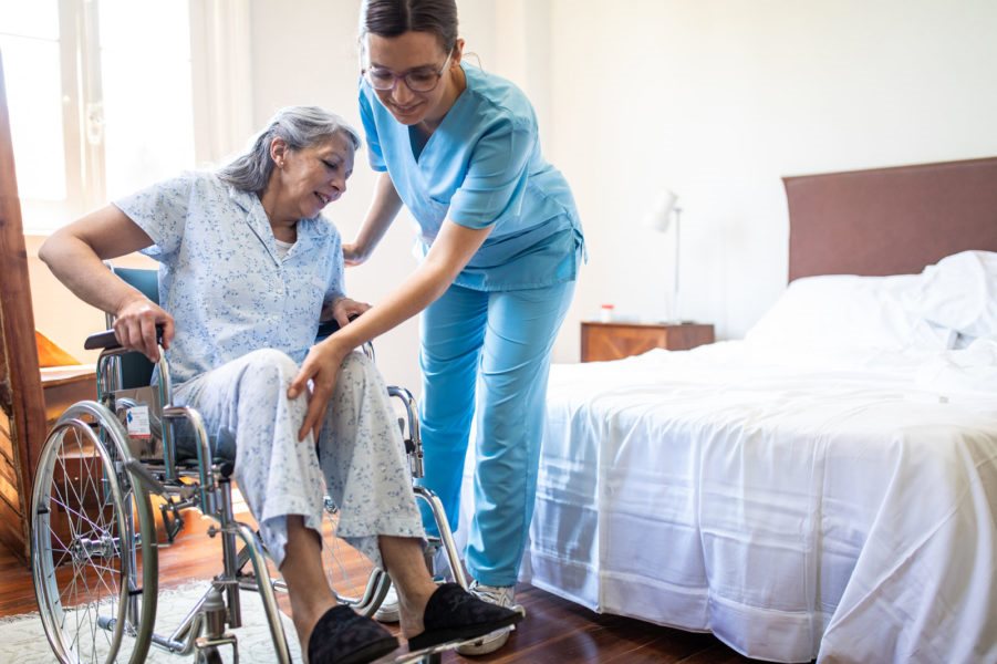 Caregiver in blue scrubs helping an elderly woman rise from a wheelchair in a home care bedroom setting