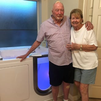 Walk In Tub Reviews Testimonials Feedback 2 Smiling senior couple standing beside their newly installed Ella walk-in tub with blue chromotherapy lighting