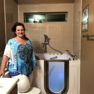 Walk In Tub Reviews Testimonials Feedback 3 Smiling woman in blue floral top posing beside her installed Ella walk-in tub with side door entry in a tiled bathroom