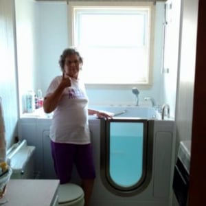 Walk In Tub Reviews Testimonials Feedback 49 Senior woman giving a thumbs up next to her filled walk-in tub with door open in a bright bathroom