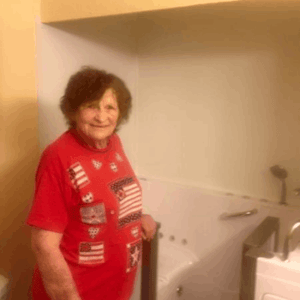 Walk In Tub Reviews Testimonials Feedback 63 Senior woman in red shirt smiling proudly beside her newly installed Ella walk-in tub with grab bar in bathroom