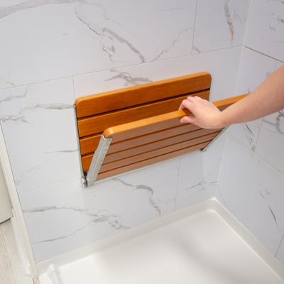 Hand folding down a teak wood wall-mounted folding shower seat with stainless steel brackets in an accessible shower stall