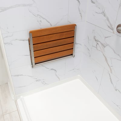 Teak wood folding shower seat mounted open on marble wall panel above an accessible shower stall base