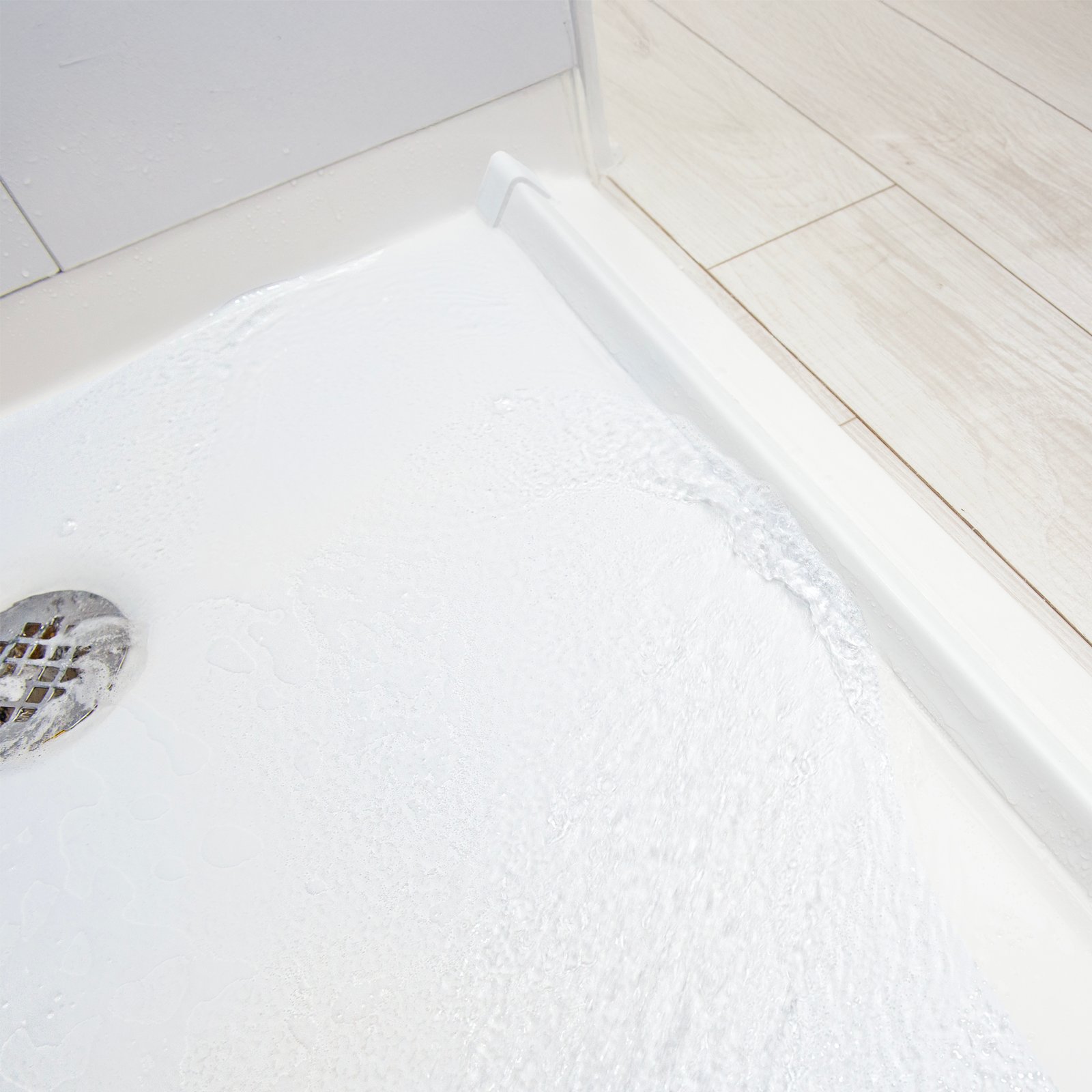 Collapsible Rubber Dam for Barrier Free Shower Bases - Close-up of water draining on a white accessible shower base with a rubber dam threshold next to light wood-look tile flooring