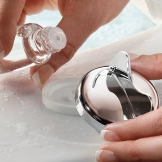 Manos vertiendo aceite esencial en un dispensador de aromaterapia cromado montado en la superficie de una bañera de acceso.