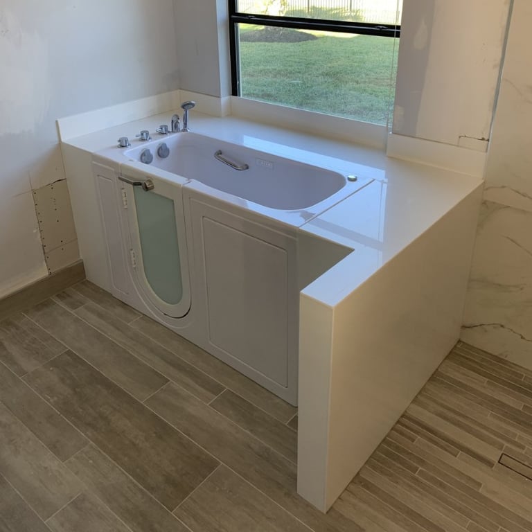 Ella walk-in tub installed in a white surround enclosure with wood-look tile flooring and a garden view window
