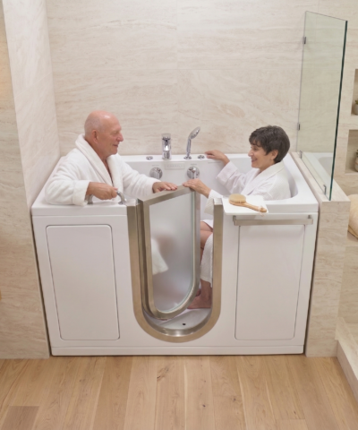 Pareja de mayores con albornoces blancos sentada junta y sonriendo dentro de una bañera de acceso Ella Companion con dos asientos y puerta de acero inoxidable